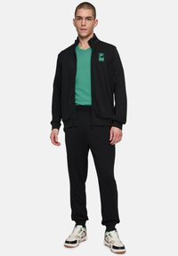 Veste noire zippée avec col haut, chemise turquoise en dessous, pantalon de survêtement noir et chaussures de sport avec des accents verts. Logo Fila sur la veste.