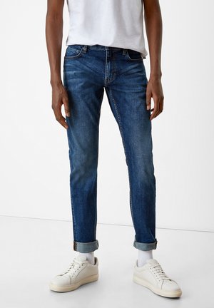 Hombre vestido con jeans de mezclilla azul puño doblado, calcetines blancos, zapatillas blancas y una camiseta blanca, de pie frente a un fondo blanco liso.