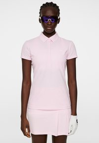 Jasnoróżowa koszulka polo z kołnierzykiem i krótkimi rękawami, w połączeniu z dopasowaną plisowaną spódnicą. Biały detal z logo na piersi.