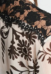 Schwarze SpitzenDetails und florale Muster auf einer cremefarbenen Bluse; mit strukturiertem schwarzem Spitzenstoff an den Schultern und fließenden Ärmeln.