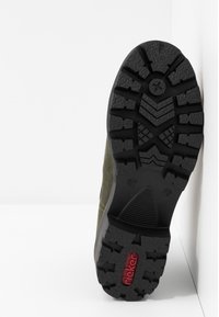 Semelle d'une botte en caoutchouc noire avec des motifs de semelle profonds et un logo rouge "rieker" près du talon, incliné contre un mur blanc.