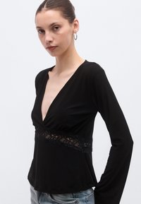 Top negru cu mâneci lungi, decolteu în V adânc și accent din dantelă în talie. Textură netedă, croială mulată. Purtați cu blugi deschiși la culoare.