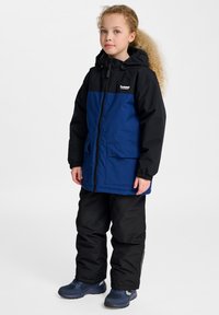 Veste imperméable bicolore noire et bleue avec capuche, fermeture éclair à l'avant et deux grandes poches. Associée à un pantalon isolant noir et des chaussures bleues.