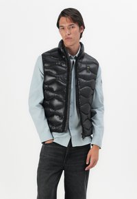 Gilet imbottito nero con finitura lucida, caratterizzato da un colletto alto, chiusura con zip e un design leggero, indossato sopra una camicia azzurra chiara.
