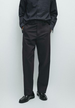 Mężczyzna ubrany w czarne, dopasowane spodnie, czarne skórzane mokasyny oraz ciemną, kraciastą koszulę z długim rękawem, z rękami w kieszeniach.