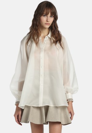 Žena s dlhými hnedými vlasmi nosí priehľadnú bielu blúzku na gombíky s nafúknutými rukávmi a béžovú rozšírenú sukňu, stojaca pred jednoliatym pozadím.