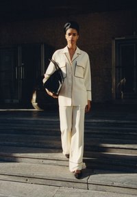 Ensemble sur mesure couleur crème avec des passepoils noirs contrastants, comprenant une chemise à boutons et un pantalon large. Tient un sac noir.