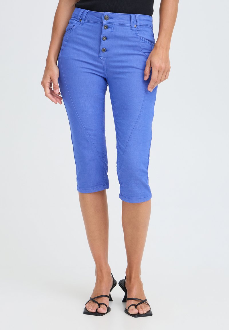 Une femme porte un pantalon court bleu vif avec quatre boutons et des talons noirs ouverts à l'avant avec des brides, debout devant un fond clair uni.