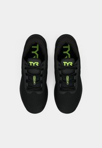 Zapatillas deportivas negras con parte superior de malla, que presentan un logo y detalles en verde, punta redonda y cordones. Diseñadas para la comodidad y flexibilidad.