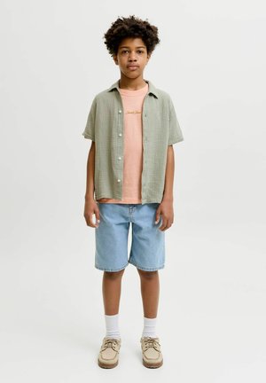 Garçon debout, portant une chemise verte claire à boutons par-dessus un t-shirt rose, un short en jean, des chaussettes blanches et des chaussures beiges, sur un fond blanc uni.