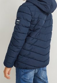 Granatowa kurtka puchowa z poziomymi przeszyciami i kapturem. Posiada naszywkę na lewym ramieniu, zestawiona z niebieskimi dżinsami.