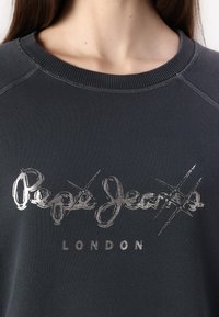 Felpa grigia scura con scollatura a costine, caratterizzata da un logo metallizzato color argento "Pepe Jeans LONDON" e dettagli a croce sotto il testo.