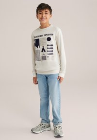 Světle šedá mikina s náměstími modrými grafikami, modré džíny a šedé tenisky. Mikina obsahuje různé vzory a text "BLUE RIDGE EXPLORER."