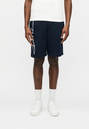 Hombre con pantalones cortos azul marino con letras blancas en el lado izquierdo, calcetines blancos hasta el tobillo, zapatillas blancas y camiseta blanca, de pie frente a un fondo liso.