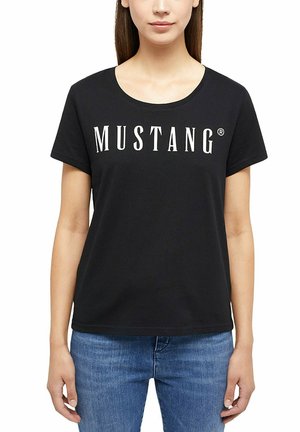 Donna che indossa una t-shirt nera a maniche corte con la scritta "MUSTANG" bianca sul petto e jeans blu, in piedi davanti a uno sfondo bianco.