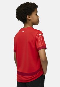 Punane spordijerseys, millel on lühikesed varrukad, tekstureeritud õlad ja ühtlane tagaosa. Sisaldab kontrastset ribaga krae ja põhiaktsentide ääriseid.