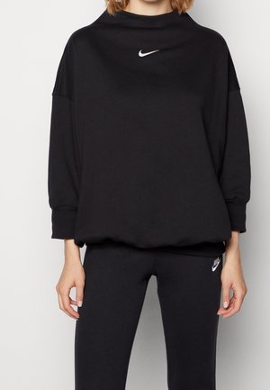 Vrouw draagt een zwarte Nike-trui met een wit logo op de borst en bijpassende zwarte leggings met een logo op de linker dij.