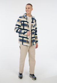 Jonge man die glimlacht, gekleed in een gestreepte blauwe, beige en witte jas, beige broek en blauwe en zwarte sneakers tegen een witte achtergrond.