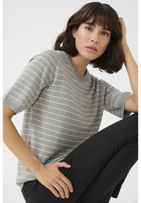 Vrouw met schouderlang donker haar, een grijze, gestreepte, korte mouw trui en zwarte broek, zittend met een hand op haar hoofd en haar knie.