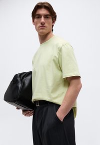 Νέος άνδρας με ανοιχτό κίτρινο μπλουζάκι και μαύρο παντελόνι κρατά μαύρη δερμάτινη τσάντα με το ένα χέρι, ενώ το άλλο χέρι είναι στην τσέπη, με φόντο λευκό.