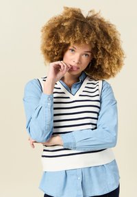 V-Ausschnitt, ärmelloser Pullover in Weiß mit marineblauen Streifen, über einem hellblauen Hemd mit Kragen getragen. Strukturierter Strickstoff mit lässiger Passform.