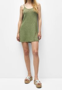 Robe en maille verte à fines bretelles, longueur mi-cuisse, coupe décontractée. Associée à des sandales à plateforme blanches ornées de motifs floraux.