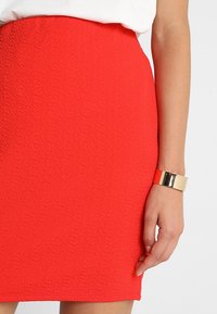 Červená přiléhavá sukně s texturovaným materiálem, vysokým pasem a hladkým lemem. Ruka zdobená lesklým zlatým náramkem.