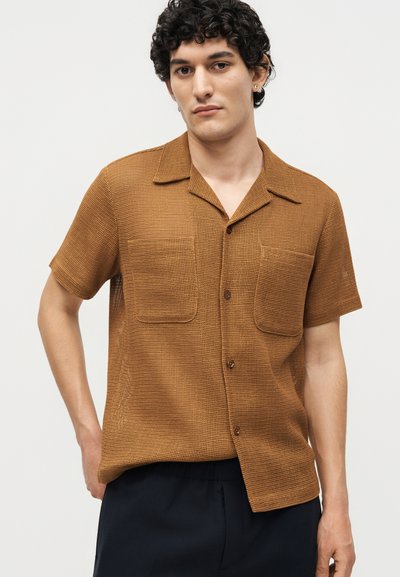 Νεαρός άνδρας με κατσαρά μαλλιά που φοράει καφέ υφασμάτινο κοντομάνικο πουκάμισο με κουμπιά και σκούρο παντελόνι σε απλό φόντο.