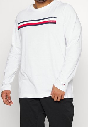 Mann trägt ein weißes Langarmshirt von Tommy Hilfiger mit blau-roten Streifen und Schriftzug, kombiniert mit schwarzer Hose, gezeigt vom Hals bis zur Hüfte.