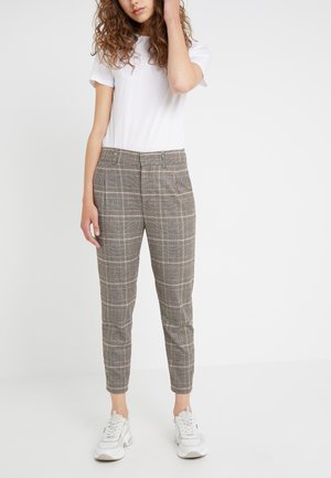 Woman wearing white t-shirt, gray plaid tapered pants, and white sneakers, standing against plain light background.