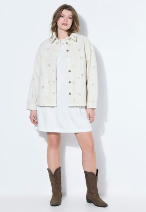 Femme portant une robe blanche, une veste crème avec des motifs de cœurs, et des bottes de cowboy marron, debout devant un fond clair uni.