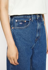 Džínové kalhoty v tmavě modré barvě s vysokým pasem a designem se pěti kapsami. Obsahují měděné nýty a malou štítkovou etiketu na přední kapse.