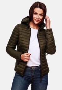 Veste rembourrée vert olive avec capuche, présentant une texture matelassée, une fermeture éclair à l'avant et des poches latérales, portée sur une chemise blanche et un jean bleu.