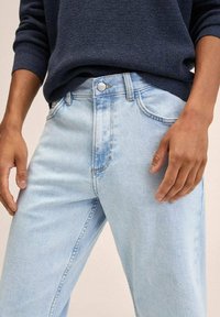 Vaqueros de mezclilla azul claro con un ligero desgaste, diseño de cinco bolsillos y cierre de botón. Combinados con un suéter de punto oscuro.