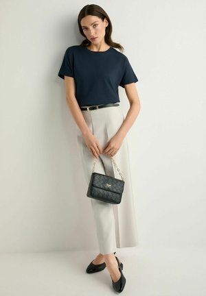 Kobieta ubrana w granatowy t-shirt, jasnobeżowe spodnie z wysokim stanem i czarnym paskiem, trzymająca czarną pikowaną torebkę na złotym łańcuszku.