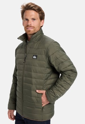 Homme portant une veste matelassée olive avec des poches zippées, se tenant contre un fond blanc, la main dans la poche.