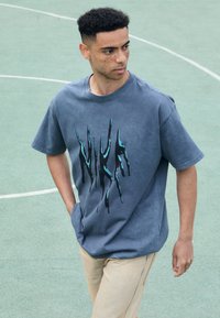 Pilka medvilnės marškinėliai su juodu ir petrolio mėlynumo abstrakčiu Nike logo, trumpomis rankovėmis, laisvo kirpimo, derinami su smėlio spalvos kelnėmis. Lygus paviršius.