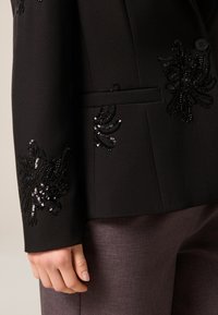 Zwarte getailleerde jas met fijne zwarte kralen- en paillettenborduursels, een gladde stoftextuur en een enkele knoopsluiting.