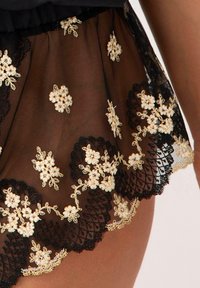 Czarna przejrzysta tkanina z złotym haftem w kwiaty i koronkowym wykończeniem, prezentująca delikatny wzór i falowane krawędzie u dołu.