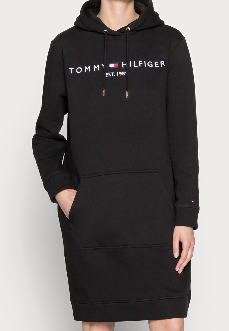 Schwarzes Hoodie-Kleid mit vorderer Kängurutasche, Kordelzugkapuze und weißem Logodruck. Hergestellt aus weichem Baumwollmix.