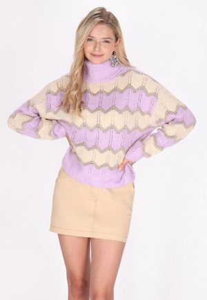 Pull en tricot avec motif en chevron en lilas et crème, avec un col montant, une coupe ample et des poignets côtelés. Le modèle porte également une jupe beige.