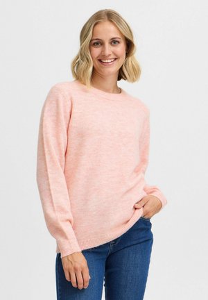 Un pull rose clair en matériau doux, avec de longues manches et un ourlet côtelé. Porté de manière décontractée avec un jean bleu foncé.