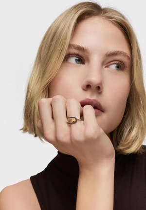 Mujer joven con cabello rubio corto apoyando la barbilla sobre la mano, lleva una blusa sin mangas negra y un anillo rectangular dorado y negro.