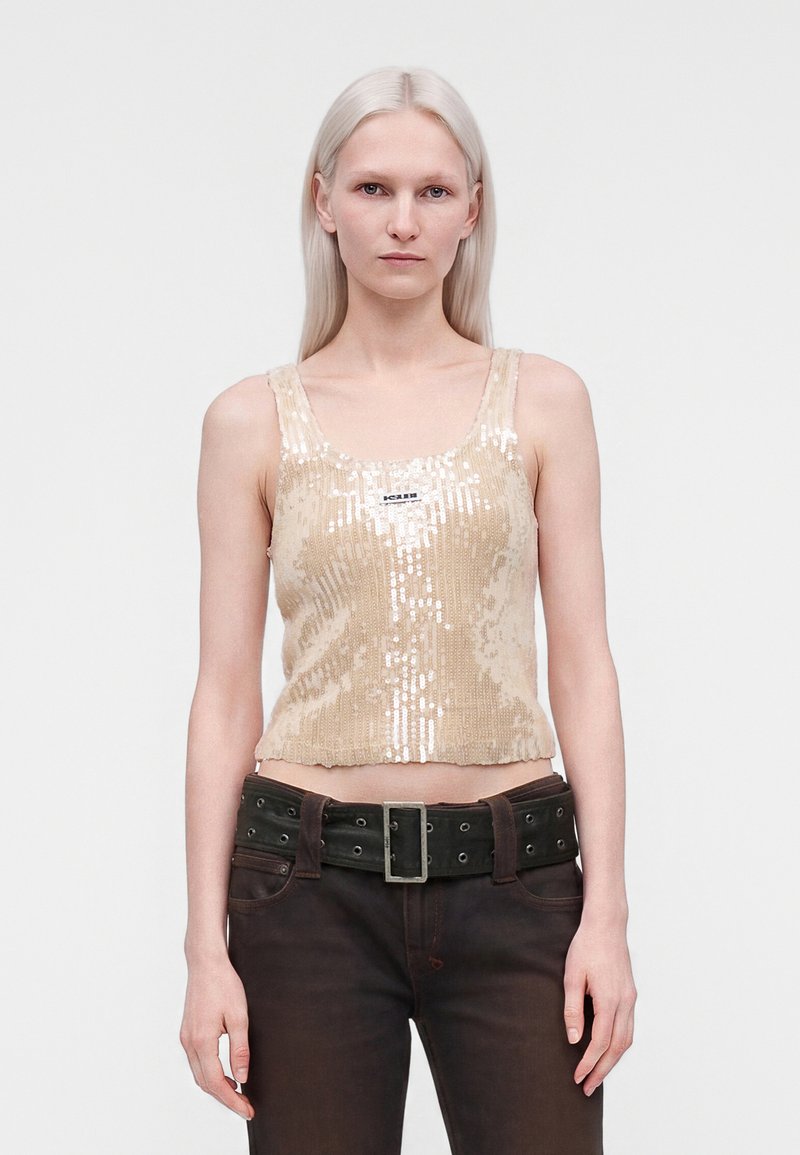 Jeune femme aux longs cheveux blonds clairs portant un haut beige sans manches à sequins et un pantalon marron foncé avec une large ceinture.