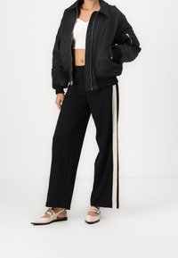 Osoba nosząca czarną kurtkę bomber, białą krótką bluzkę, czarne szerokie spodnie z białym paskiem po boku oraz beżowe czółenka z paskami.