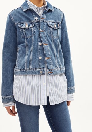 Persoon draagt een blauwe spijkerjas met zilveren knopen over een lichtblauw-wit gestreept blouse met knopen en een blauwe spijkerbroek.