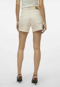 Pantalones cortos de mezclilla color crema con una cintura alta, costuras visibles y dos bolsillos traseros. Llevados con sandalias de tacón marrones.