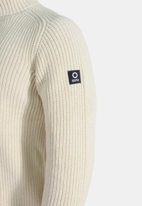 Maglione lavorato a maglia di colore crema, caratterizzato da una texture a coste, collo alto e un piccolo etichetta nera con testo bianco sulla manica sinistra.