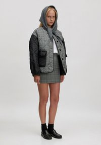 Gestepptes Bomberjackett in Schwarz und Grau mit Karo-Design, taillierter grauer Karorock, grauer Schal, schwarze Loafer mit Fransenakzenten und schwarze Socken.