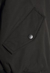 Černá bunda s šikmou kapsou a kulatým černým cvokem, hladkým textilním povrchem a kontrastním žebrovaným lemem.